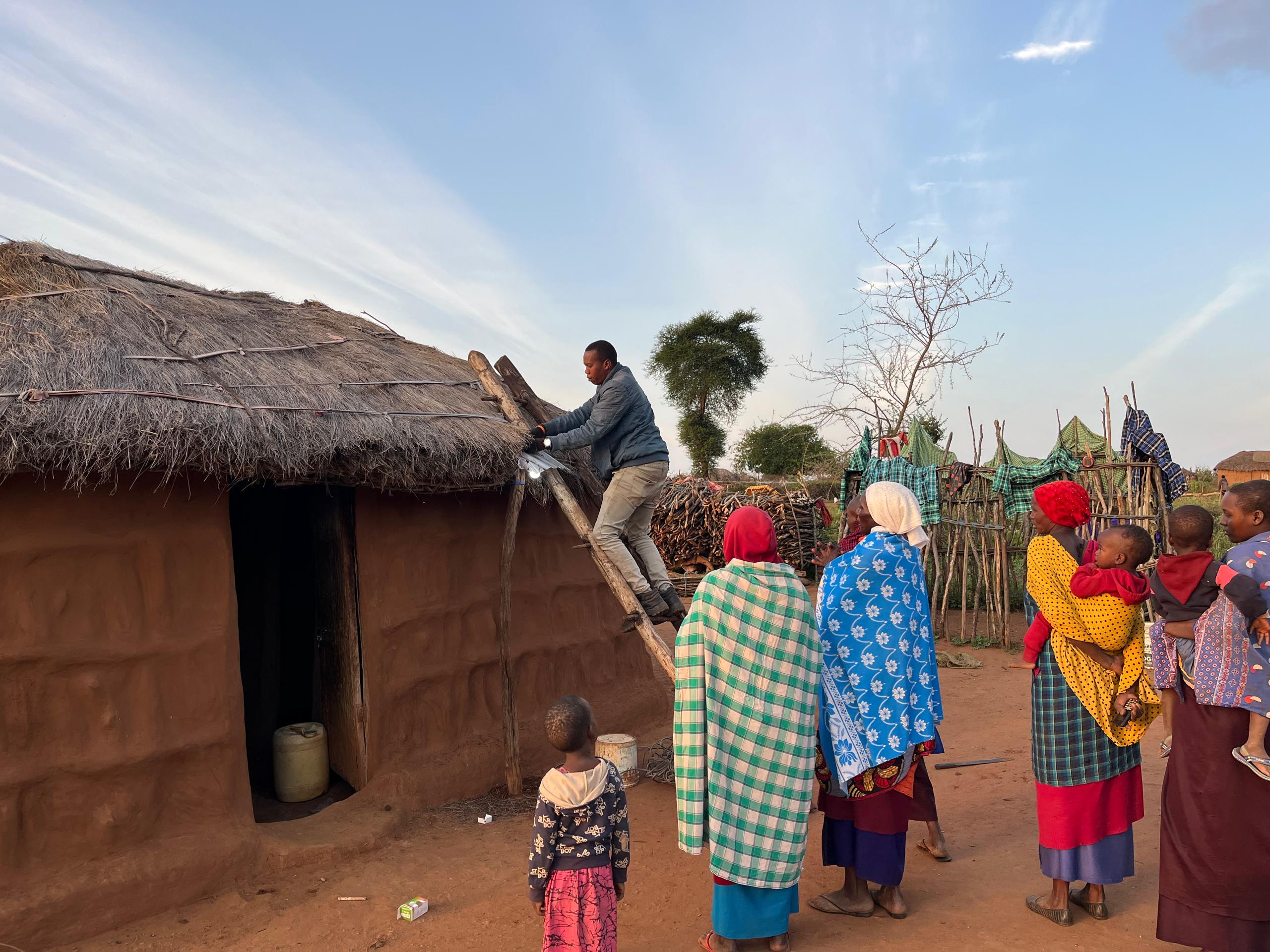 A group of women and chidren in traditional Maasai clotes watch as our staff connects a light to their home.